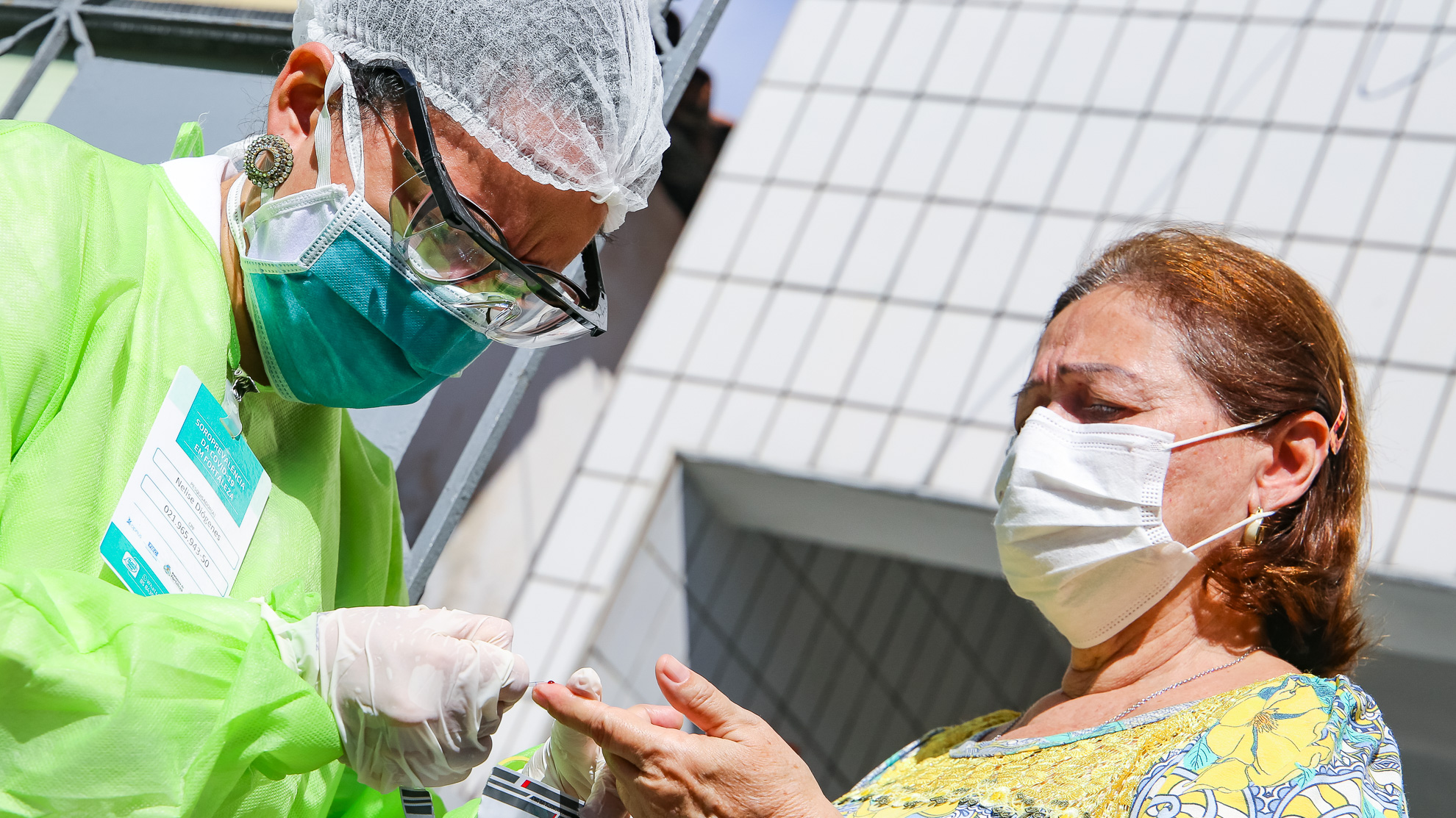 mulher profissional de saúde com máscara, touca e óculos aplicando teste em dedo de senhor que usa máscara